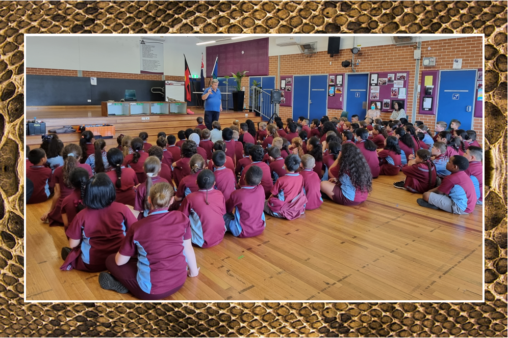 A large group of students sitting on the floor in the hall listening to the snake handler.